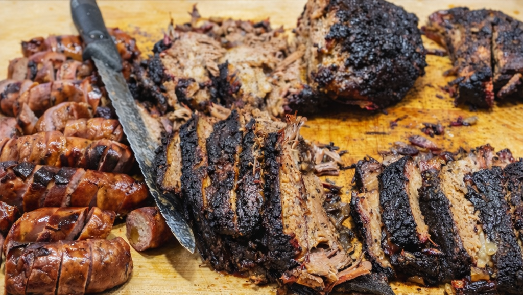 Wooden cutting board with sliced barbecue meats and sides on a wooden table