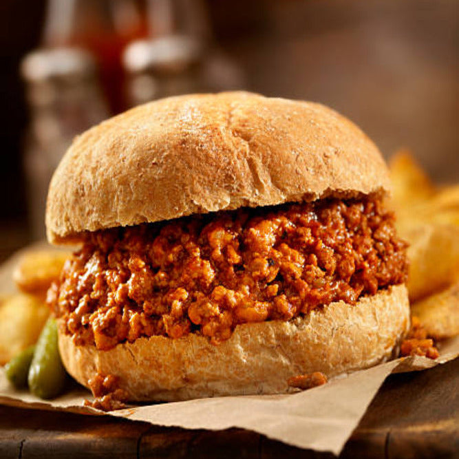 Sloppy joe sandwich on a bun with fries and a bottle of ketchup in the background.